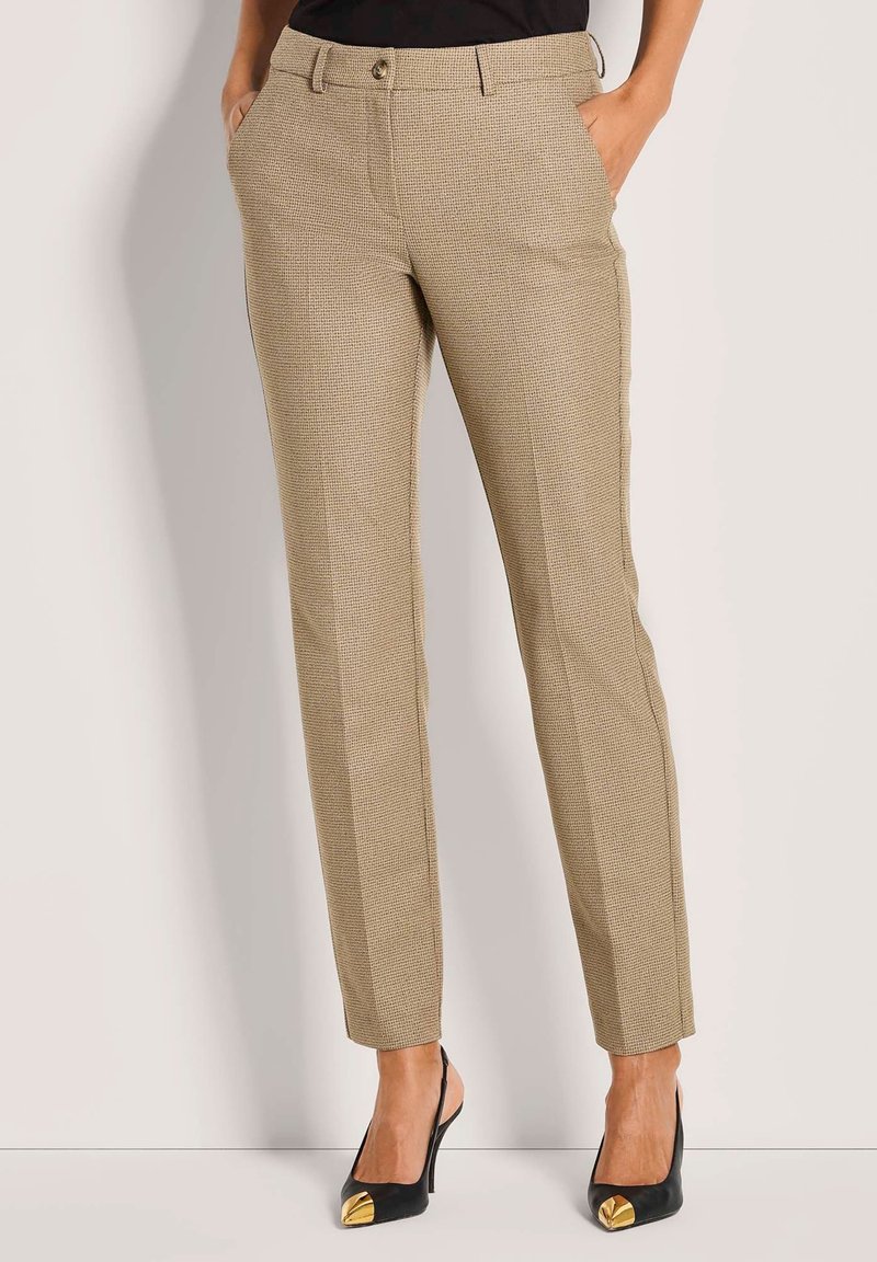 Vrouw in beige slim-fit geblokte broek met zakken en zwarte hoge hakken met gouden tenen, staand met handen in de zakken.