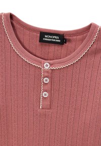 Haut tricot à côtes rose, avec un col rond, agrémenté d'une bordure festonnée blanche et de trois boutons décoratifs au niveau de la patte de boutonnage. Étiquette de marque visible.