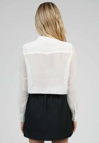 Femme aux longs cheveux blonds portant une blouse blanche transparente à manches longues rentrée dans une jupe noire taille haute, de dos sur un fond gris.