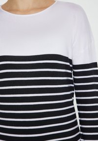 Tricou cu dungi alb-negru, cu mâneci lungi și decolteu rotund, având partea superioară complet albă și dungi orizontale alternând alb-negru în partea de jos.