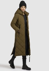 Oliven grøn, quiltet lang jakke med hætte, to sidelommelygter og sorte detaljer, parret med sorte knæhøje støvler og en strikket hue.