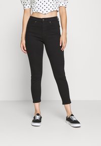 Svarta cropped jeans med slim fit, med en klassisk femficksdesign och metalldekor, kombinerade med svarta sneakers och en prickig topp.