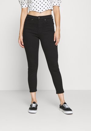 Jean noir coupé avec une coupe slim, featuring un design classique à cinq poches et une fermeture à bouton en métal, porté avec des baskets noires et un haut à pois.