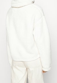 Biała polarowa bluza z wysokim kołnierzem, opuszczonymi ramionami i miękką teksturą. Charakteryzuje się krótką długością i ściągaczami przy mankietach.