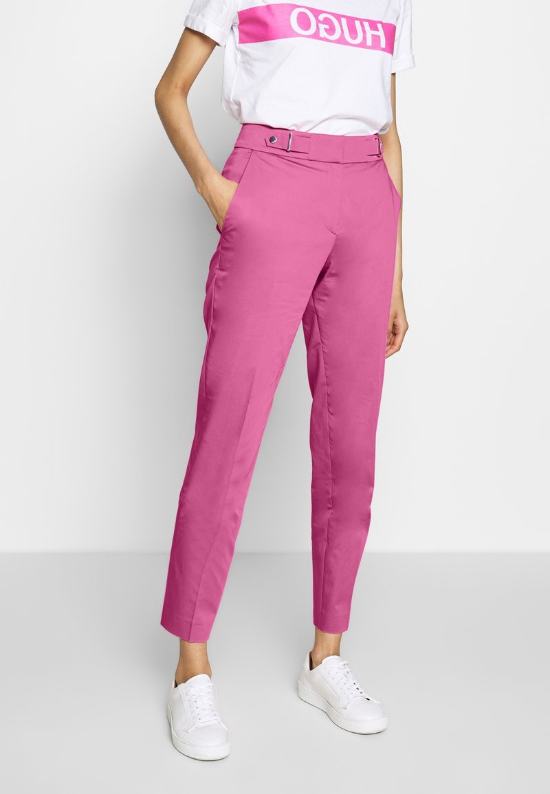 Femme portant un pantalon rose taille haute à jambes fuselées avec passants de ceinture, des baskets blanches et un T-shirt blanc avec le logo "HUGO" rose. Mains dans les poches.