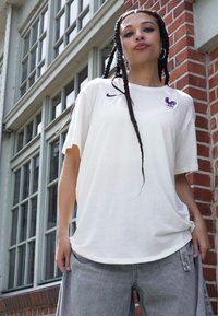 Valkoinen puuvillapaita, jossa on löysä istuvuus, varustettuna violetilla Nike-logolla ja violetilla kukko-embleemillä rinnassa. Yhdistetty harmaisiin housuihin.