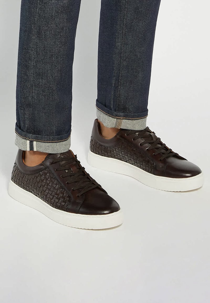 Brown leather sneakers with woven texture on sides, round toe, contrasting white rubber sole, and brown laces. Worn with cuffed dark jeans.