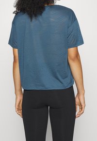 Szmaragdowa, krótka koszulka sportowa o luźnym kroju, z teksturą falistą i zaokrąglonym dołem, w zestawie z czarnymi legginsami.