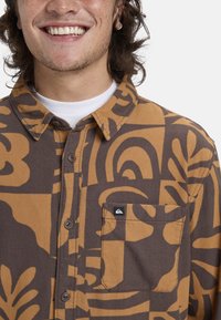Brun og gul mønstret skjorte med knapper, inkluderende en lomme med lap og logo, med en klassisk krave og blød tekstur.