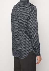 Černá košile s bílým geometrickým vzorem, dlouhými rukávy a límečkem. Materiál má hladkou texturu a uvolněný střih.