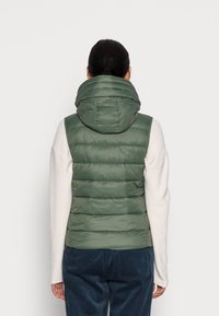 Groene gewatteerde bodywarmer met capuchon en doorgestikte delen. Materiaal heeft een gladde, glanzende afwerking. Draagt over een lichtgekleurde trui.