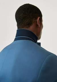 Blaues Jackett mit einem Kragen, der eine marineblaue Unterseite und hellblaue Nähte aufweist, glatte Textur, taillierte Passform und strukturiertes Design.