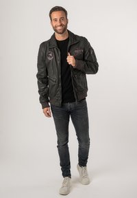 Schwarze Lederbomberjacke mit gerippten Bündchen, vorderen Taschen und gestickten Patches. Getragen über einem schwarzen Hemd und kombiniert mit dunklen Jeans und weißen Sneakern.