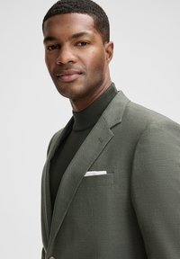 Giacca verde oliva con tessuto testurizzato, chiusura con un bottone singolo e un fazzoletto da taschino bianco. Sotto, un dolcevita scuro aderente.