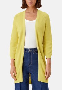 Světle žlutý pletený kardigan s otevřeným předním dílem, žebrovanou texturou a tříčtvrtečními rukávy. Kombinovaný s bílým topem a džínami s vysokým pasem.