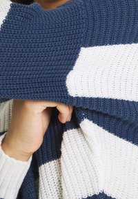 Pull en maille avec des rayures horizontales bleu marine et blanches, tissu texturé et coupe ample, une main saisissant l'ourlet.