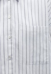 Chemise blanche avec fines rayures verticales noires, poche poitrine devant et boutons blancs le long de la patte de boutonnage.