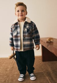 Ručīgi izšūti flaneles žakete ar vilnas apkakli, krēms krekls, tumši bikses un melni sporta apavi; turēt koka lidmašīnas rotaļlietu.