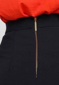 Černé kalhoty s vysokým pasem a zlatým zipem s hladkou texturou. Zip se nachází vertikálně vpředu, doplněný červenou košilí nahoře.