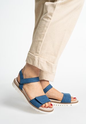 Feet wearing beige pants and blue casual sandals with adjustable straps and white soles, against a white background.