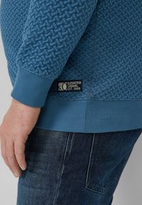 Niebieska teksturowana bluza z prążkowanym dołem i mankietami, z czarną metką z boku z napisem "S.O. Elevated Casual Est. 1969".