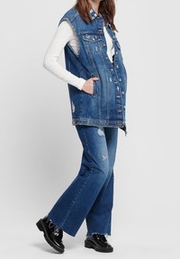 Denim ärmlös väst med framknappar och bröstfickor, bärs över en vit långärmad topp. Matchas med utsvängda jeans med slitna detaljer.