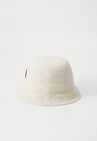 Chapeau seau beige clair en coton avec une couronne ronde et un large bord descendant. Présente un petit logo noir sur le côté.