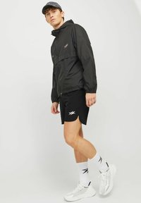 Mann in schwarzer Windjacke, schwarzen Shorts, weißen Socken, weißen Sneakers und dunklem gemustertem Cap, der vor einem einfachen hellen Hintergrund steht.