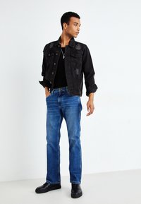 Jachetă din denim negru cu accente deteriorate, purtată peste o cămașă neagră, asortată cu blugi albaștri și adidași negri. Design simplu și casual.