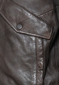 Close-up van een donkerbruine leren jas die het gestructureerde materiaal, de gedetailleerde stiksels en een drukknoop op een flapzak toont.