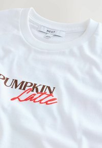 Bijela pamučna majica s natpisom "PUMPKIN Latte" u tamnosmeđoj i crvenoj boji, s okruglim izrezom i rebrastim ovratnikom.