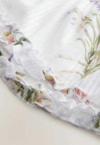 Witte stof met een bloemenpatroon aan de rand, voorzien van delicate ruffles. Zachte textuur met subtiele horizontale strepen.
