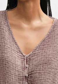Light mauve top with a textured, crinkled fabric, featuring a deep V-neckline and five circular buttons down the front.