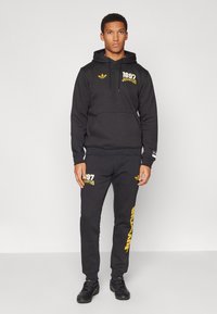 Svart bomullshoodie och sweatpants-set med gul "1897 JUVENTUS" text. Har en känguruficka och ribbade ärmslut. Standard passform.