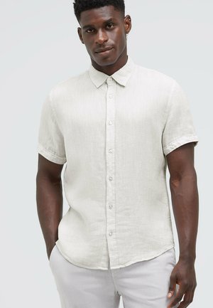 Mann trägt ein hellbeiges, kurzärmeliges Hemd mit Kragen, steht mit einer Hand in der Tasche vor einfachem Hintergrund.