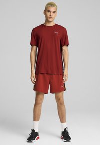 Conjunto deportivo para hombre que incluye una camiseta roja con tecnología de absorción de humedad y pantalones cortos a juego, acentuados con franjas laterales amarillas y zapatillas deportivas negras.