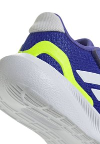 Zapatilla deportiva morada con parte superior de malla texturizada, suela blanca y un prominente acento amarillo neón en el costado. Cuenta con un talón acolchado.