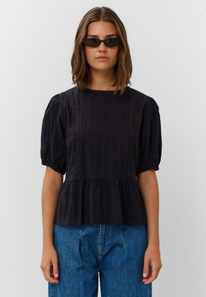 Femme portant un chemisier noir à manches bouffantes et un jean bleu, avec des cheveux courts bruns et des lunettes de soleil noires étroites, debout devant un fond uni.