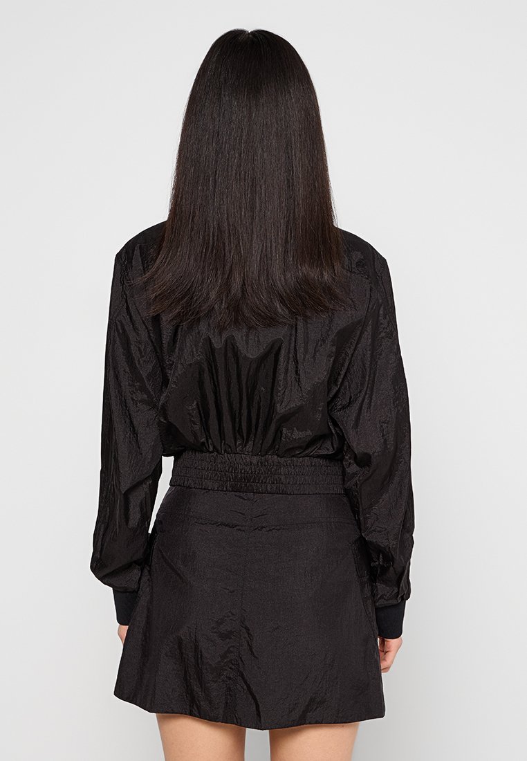 Schwarze, kurz geschnittene Jacke mit elastischem Bund, langen Ärmeln und strukturierter Oberfläche. Kombiniert mit einem passenden schwarzen Rock mit glatter Oberfläche.