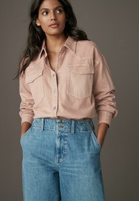 Cămașă din denim de culoare roz deschis, cu închidere prin nasturi și două buzunare pe piept, asortată cu blugi albaștri cu talie înaltă, expusă pe un fundal neutru.