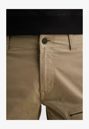 Približni prikaz bež hlača s kopčom na gumbe, omčama za remen i džepom na patentni zatvarač, nošenih s crnom majicom zašivenom unutra.