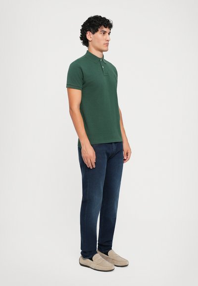 Camisa polo verde con cuello y tres botones, combinada con unos vaqueros oscuros de corte slim y zapatos tipo slip-on de color beige claro.