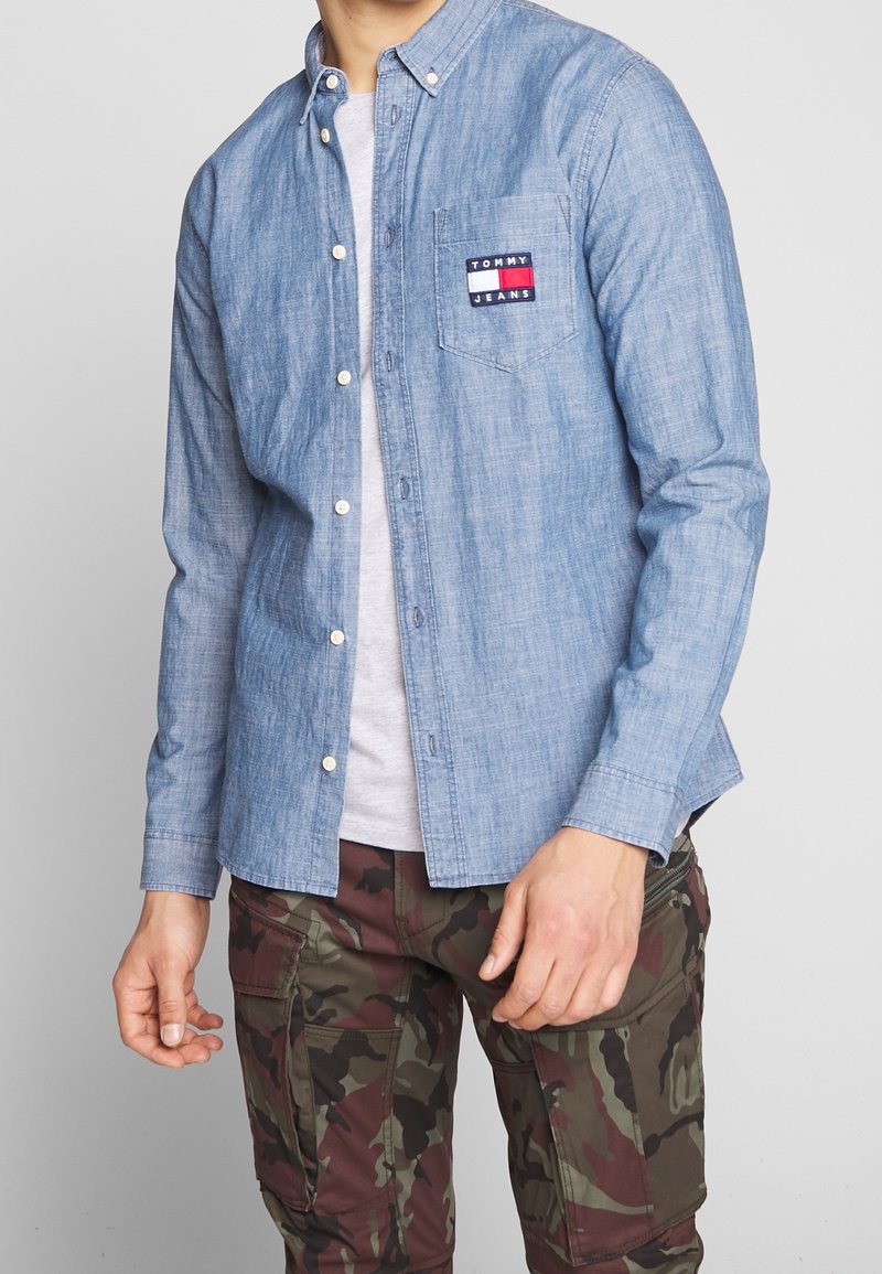 Camicia azzurra chiara in tessuto chambray con taschino sul petto con una patch del logo. Indossata sopra una maglietta grigia e pantaloni mimetici.