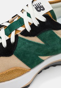 Baskets New Balance beige et vert avec lacets blancs, accents en suède noir et moutarde, et semelle blanche bordée de beige.