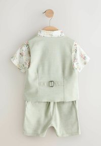 Gilet vert clair pour enfants avec sangle arrière réglable, short assorti et chemise fleurie à manches courtes suspendus sur un crochet en bois.