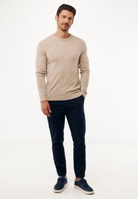 Uomo in piedi con una mano nella tasca dei pantaloni blu navy, indossando un maglione beige a maniche lunghe e sneakers blu navy su uno sfondo bianco.