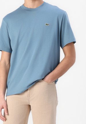 Homme portant un t-shirt bleu à manches courtes avec un petit logo crocodile vert et un pantalon beige, une main dans la poche.