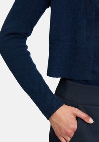 Niebieski krótki sweter z teksturowanym splotem i długimi rękawami, zaprezentowany z ciemnymi spodniami. Widoczna kieszeń i ręka lekko opierająca się na odzieży.