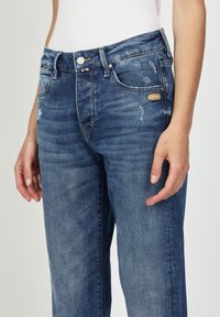Blå denimjeans med figursydd midja, lätt slitage på framsidan, femficksdesign samt metallknappar och nitar.