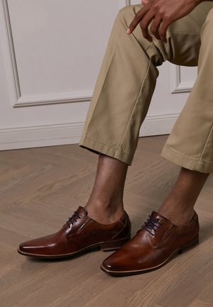 Brown leather dress shoes with pointed toes, blue laces, and decorative stitching. Worn with light beige trousers on a wooden floor.
